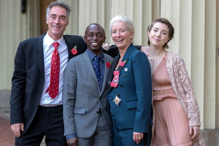 Four people stand closely, smiling under a covered walkway. They wear formal attire, including poppy pins and a medal on a blue suit.