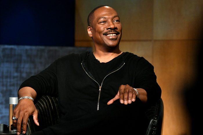 Person smiles while sitting in a black chair, against a textured background, wearing a black zip-up shirt.
