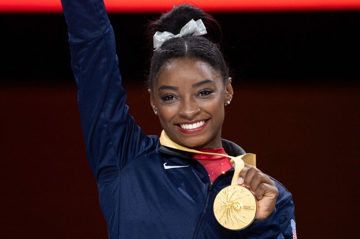A person smiles holding a gold medal against a dark background, wearing a dark jacket, with a bow in hair.