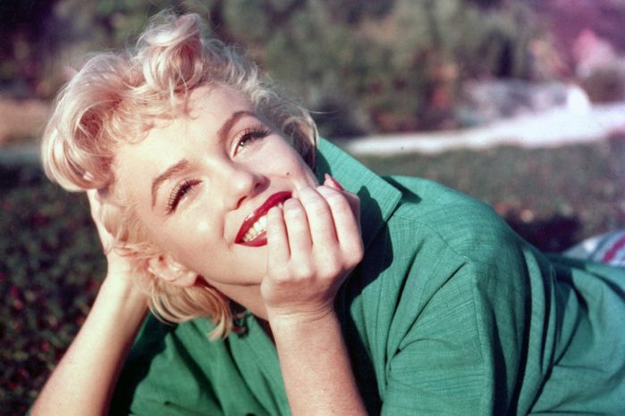 Person smiling and reclining on grass, wearing a green shirt, with a hand touching their face. Background shows blurred foliage.