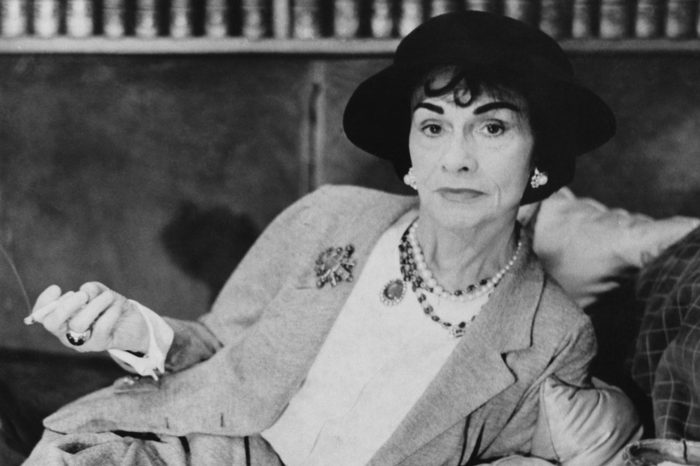 Woman reclining, holding cigarette holder, wearing elegant suit and hat, surrounded by books in a sophisticated setting.