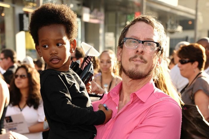 Man in pink shirt holds child with toy, amidst a crowd on a sunny day.