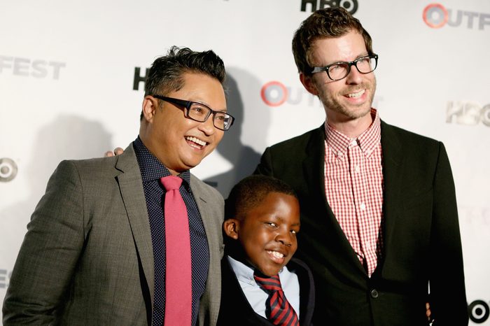 Three people smile and pose together, standing against a backdrop with "HBO" and "OUTFEST" logos, suggesting a public event or premiere.