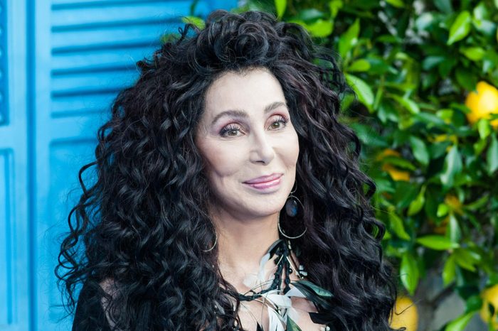 Person smiling, wearing large earrings and feather necklace, standing outdoors with blue shutters and green foliage, including lemons, in the background.