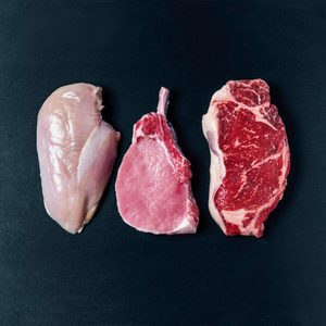 Chicken breast, pork chop, and beef steak are neatly arranged on a dark surface, showcasing various types of raw meat.