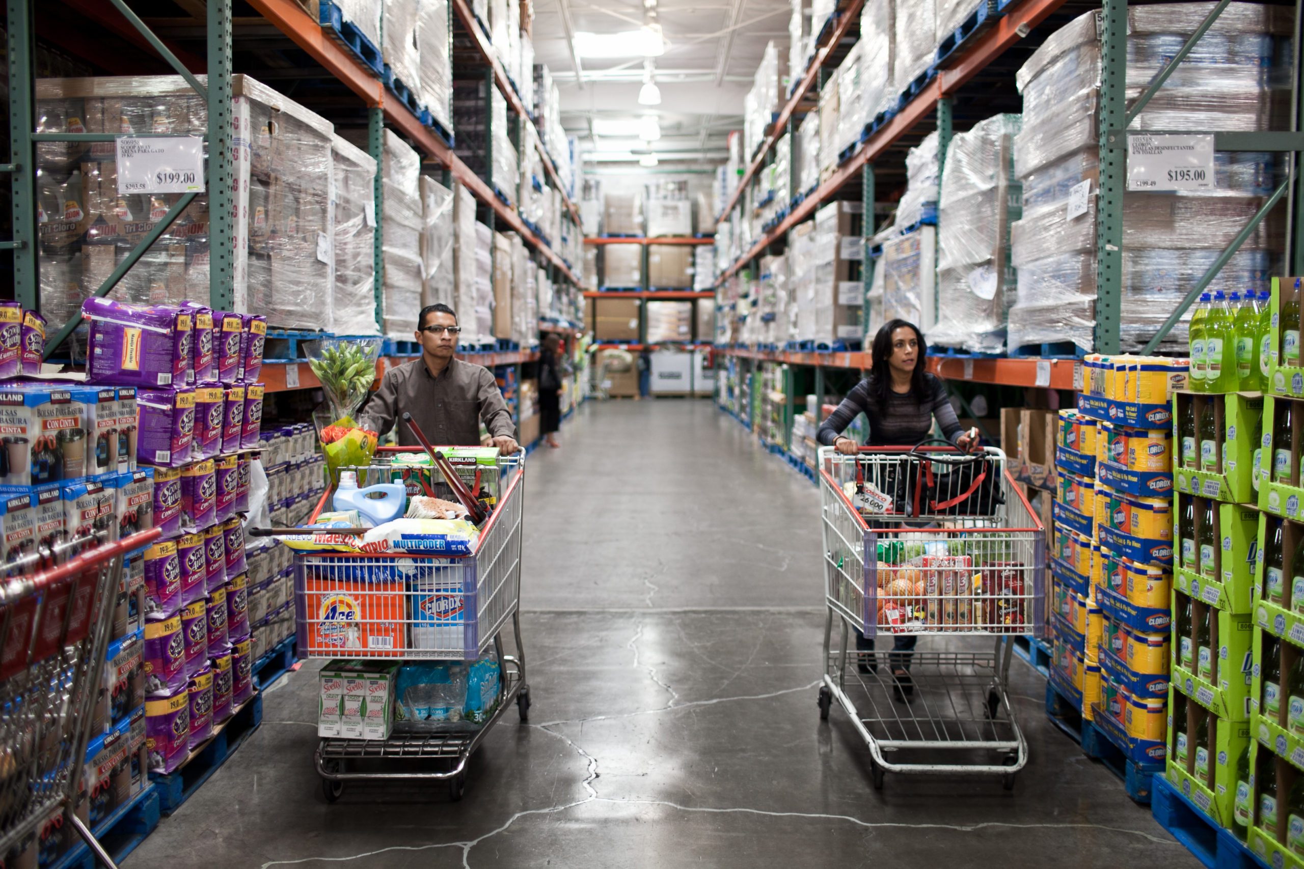 Shoppers push carts filled with groceries down an aisle in a large warehouse store, surrounded by stacked pallets and products.