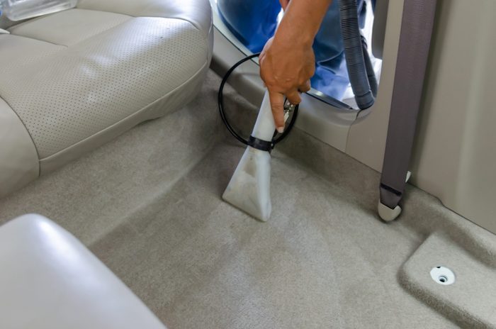 Hand operates vacuum nozzle, cleaning light-colored carpet inside vehicle near leather seat and door frame.
