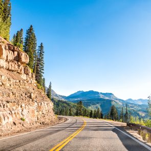 Road curves gently through mountainous landscape, bordered by rocky cliffs and pine trees, under a clear blue sky.