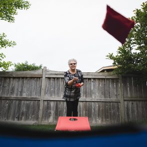 Person throws red bean bag, aiming at a hole on a raised board, outside in a fenced yard.
