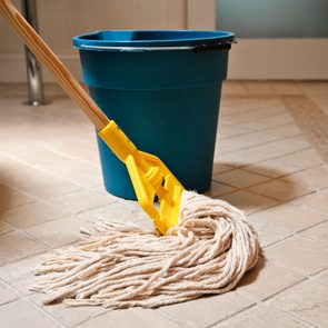 A mop rests on a tiled floor beside a blue bucket, suggesting cleaning in a household setting.