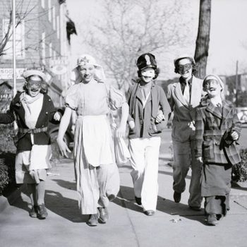Five children walk together in costumes and masks, joyfully moving along a tree-lined urban sidewalk. Sparse trees and buildings outline the street background.