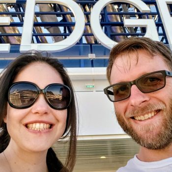 Two smiling people wearing sunglasses pose outdoors against a large "EDGE" sign in bright sunlight.