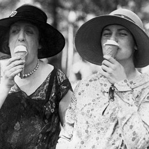 Two women enjoy ice cream cones while wearing hats in a park setting.