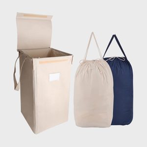 Beige laundry hamper open with handles, next to beige and navy drawstring laundry bags against a plain gray background.
