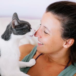 A cat licks a woman's nose while she smiles brightly, lounging indoors on a comfortable sofa.