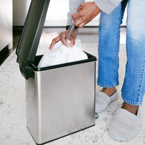 Person placing trash bag inside a stainless steel bin, wearing jeans and slippers, in a kitchen with a white rug and cabinets.