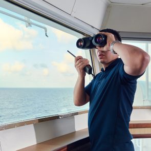 A person uses binoculars and holds a radio, observing the ocean through a ship's large window in daylight.