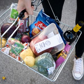 A hand holds a full shopping basket containing groceries, including vegetables, meat, snacks, and wine, on a pavement surface.