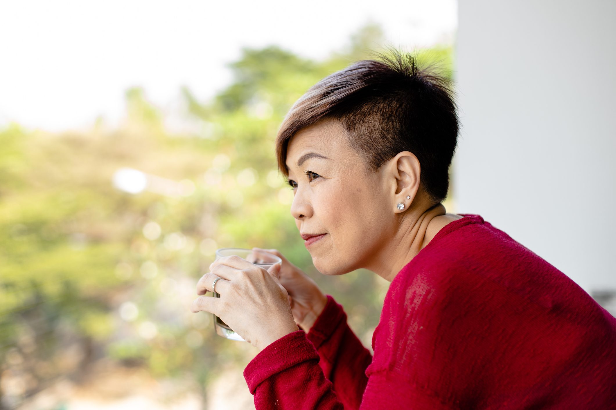 A person holds a transparent cup, gazing thoughtfully, while wearing a red sweater in a bright, blurred outdoor setting.