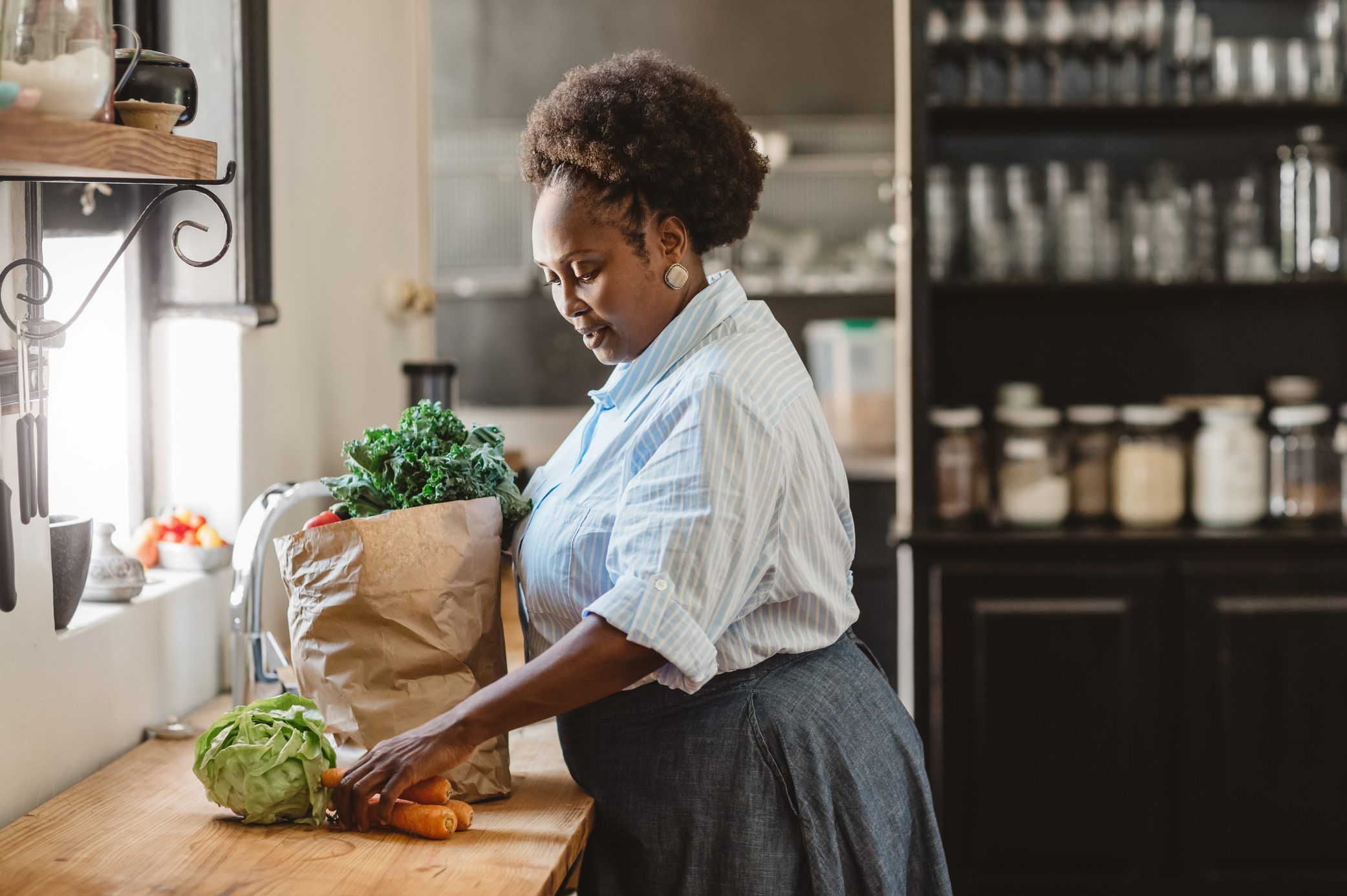 Woman unpacking groceries from a paper bag on a kitchen counter; jars and shelves visible in the background.
