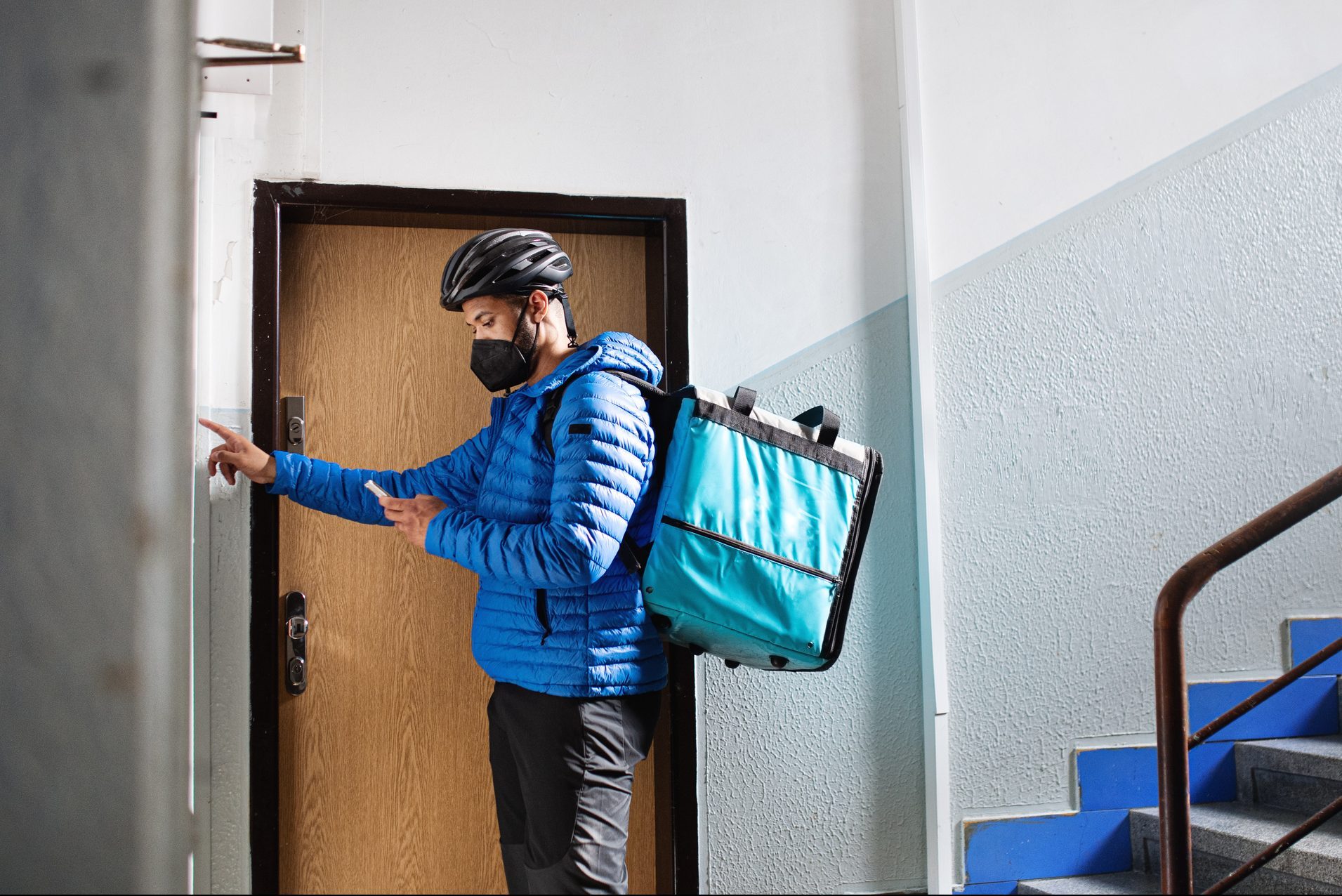 Courier wearing a helmet and mask rings a doorbell, holding a phone, in a stairwell with blue steps and a wooden door.