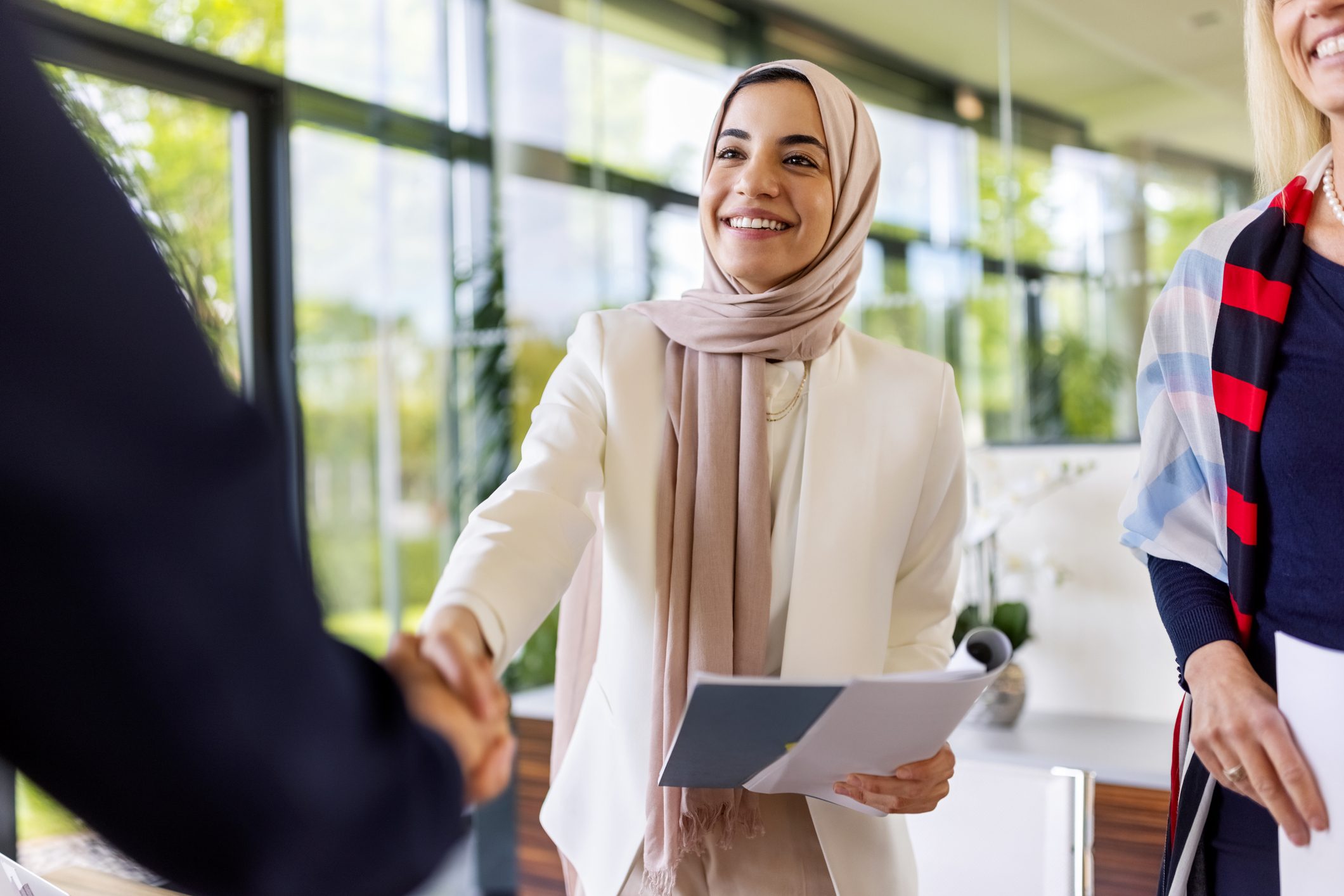 A woman in a beige hijab smiles while shaking hands in a bright office setting, holding documents.