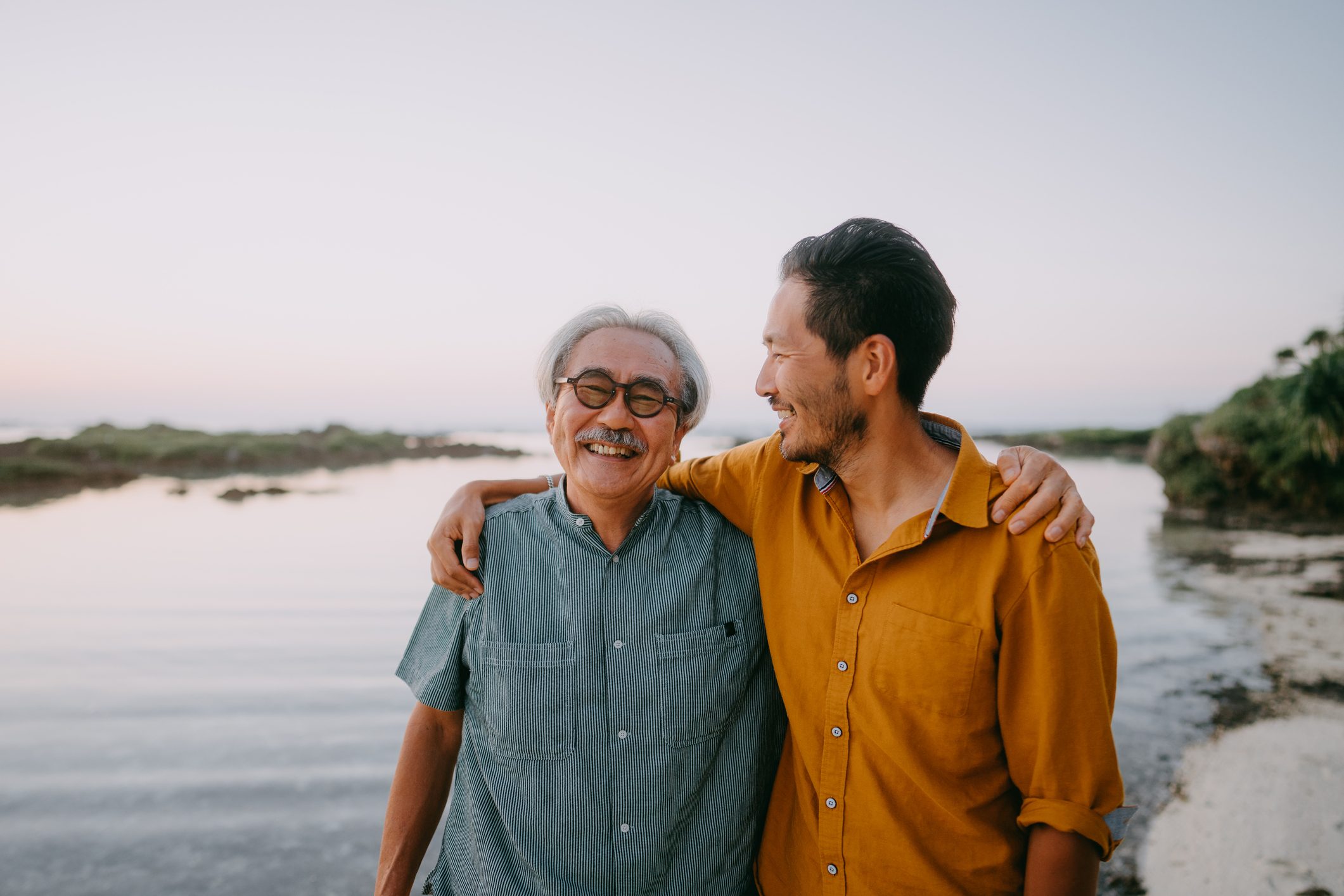 Two men smile while embracing, standing by a calm beach with a serene, pastel-colored sky in the background.