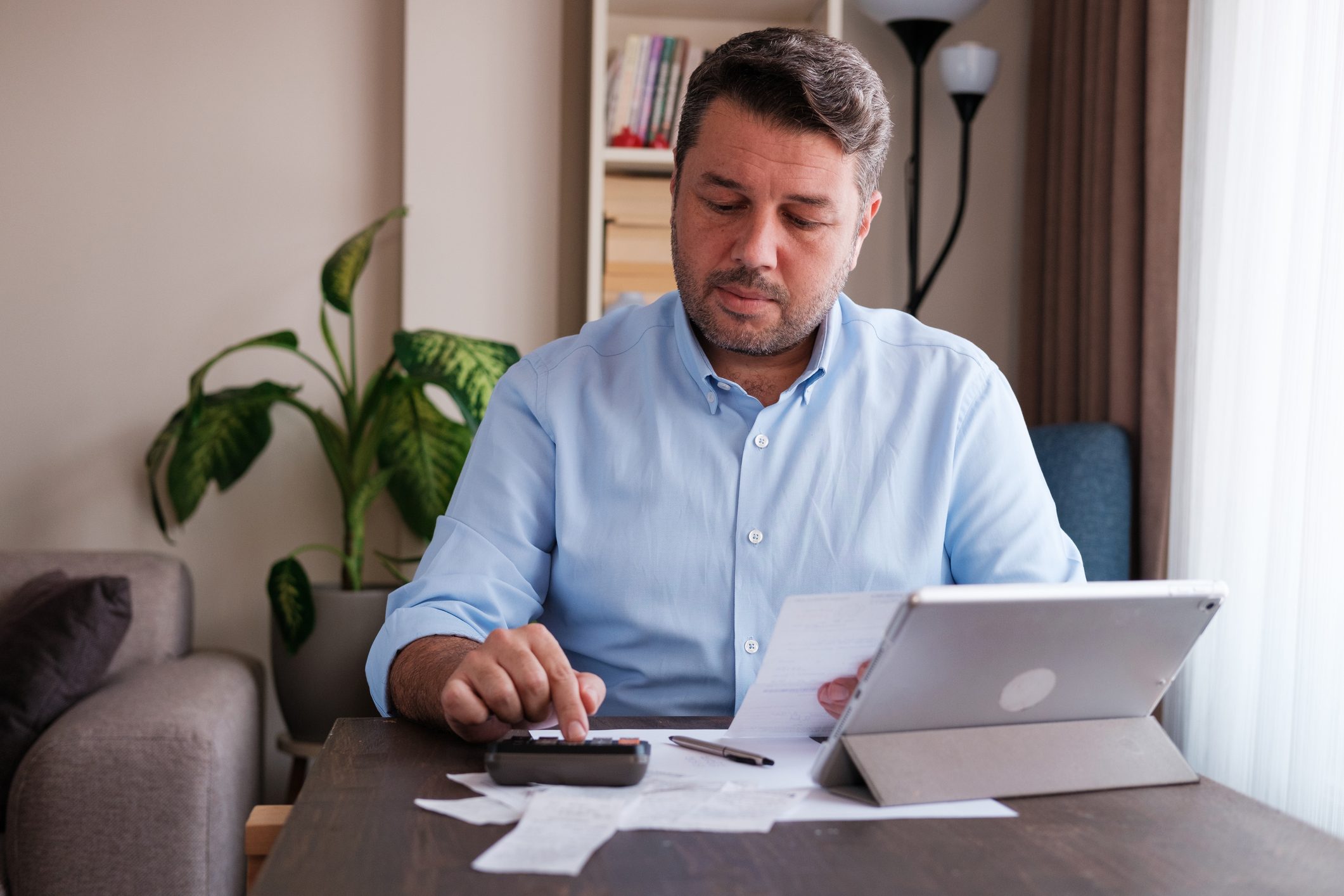 Man calculating expenses at a table, using a calculator and holding papers, surrounded by a tablet, pen, and indoor plants.