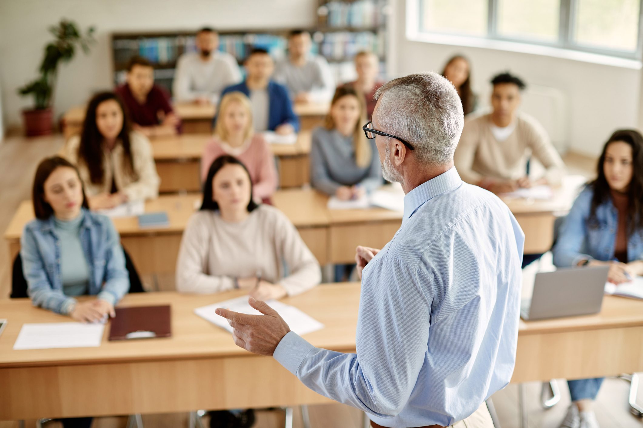 Teacher speaks to attentive students seated in a classroom, with bookshelves and windows in the background.