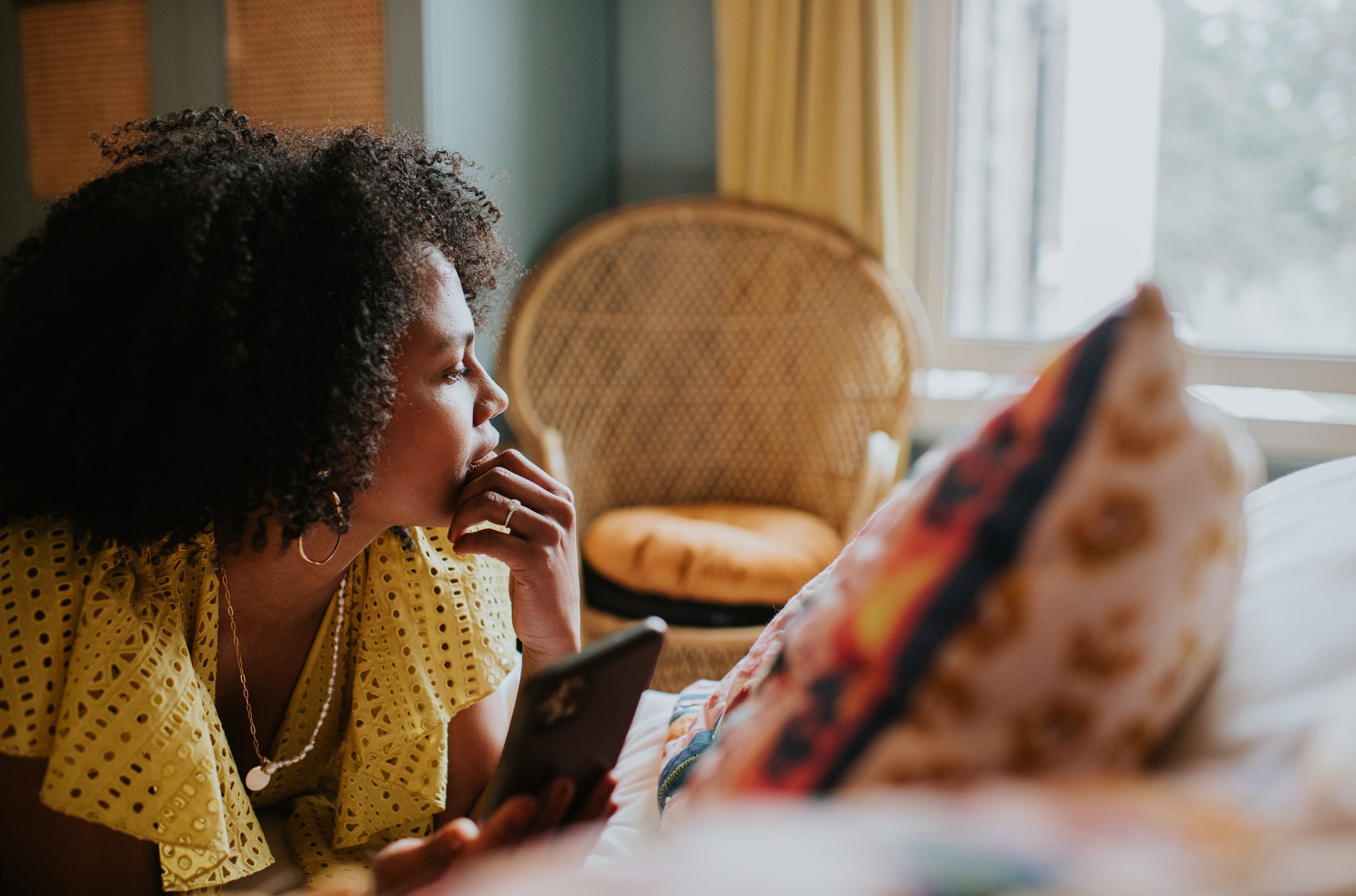A person holds a phone, lying on a bed, gazing thoughtfully toward a window in a cozy room with wicker furniture.