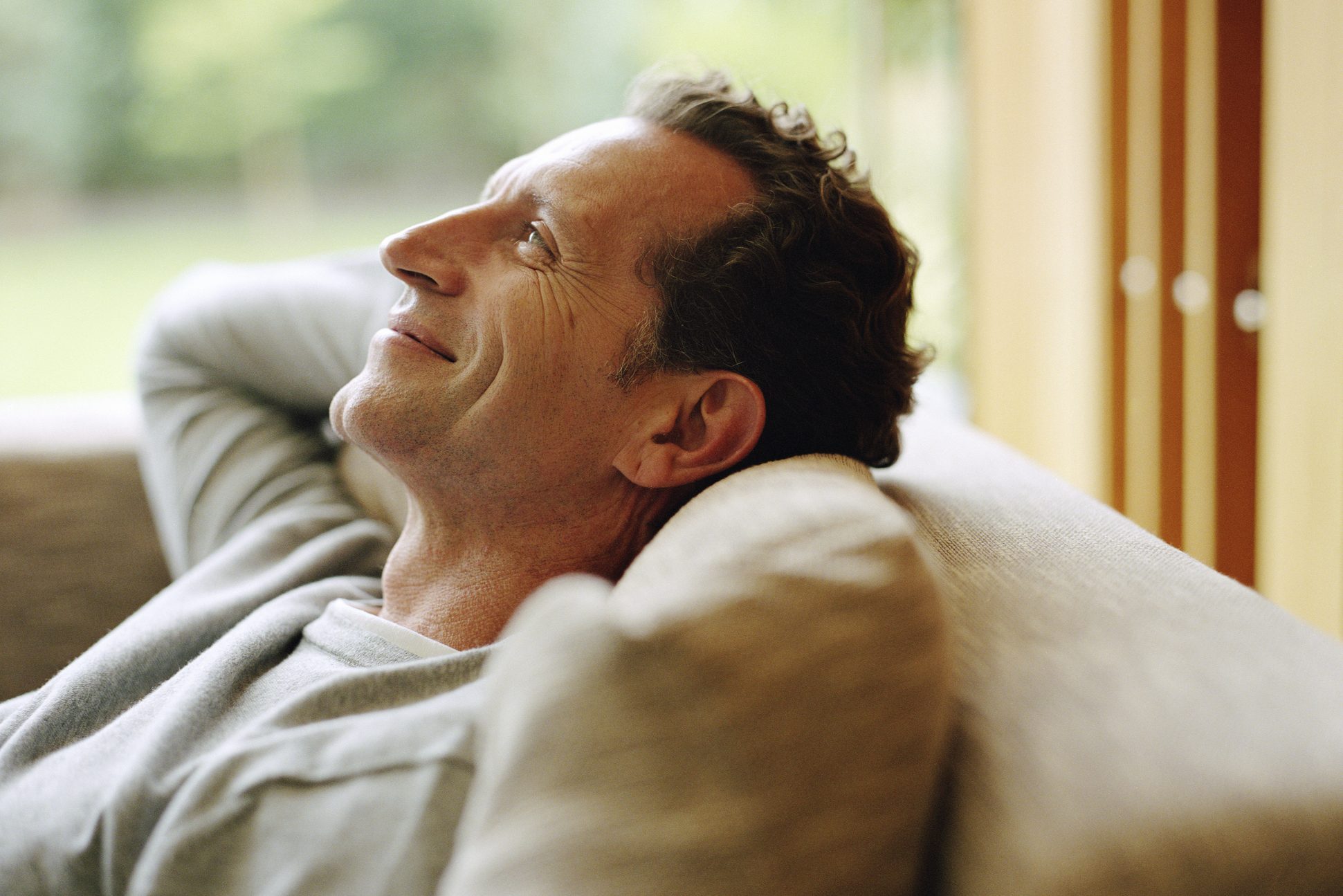Man relaxing, smiling with eyes closed, on a beige couch in a softly lit room with a blurred green garden view outside.