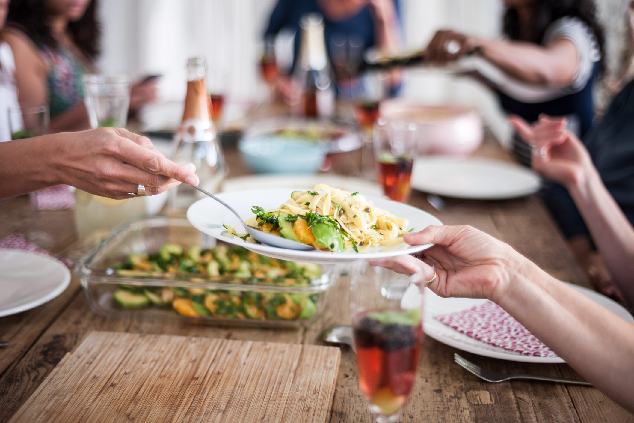 Plate of pasta is being passed at a wooden table set with drinks and other dishes, with people gathered around.