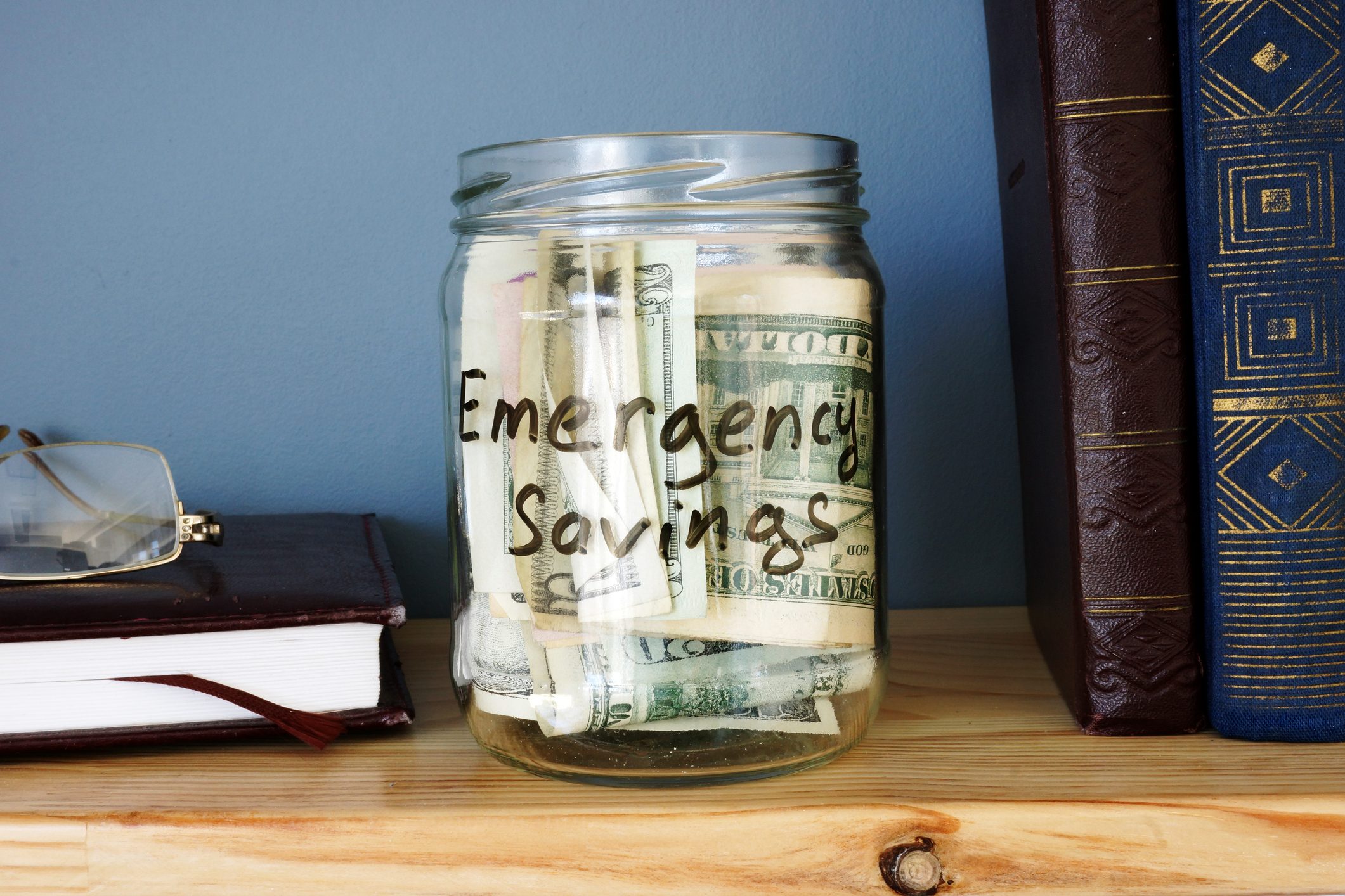 A glass jar labeled "Emergency Savings" holds money on a wooden shelf, flanked by books and eyeglasses; background is a plain blue wall.