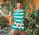 A person in a green and white striped dress stands smiling, holding a brown bag, in front of lush greenery and an orange wall.