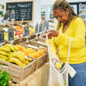 Woman selecting produce, placing fruits into a mesh bag, in a grocery store with various fruits displayed in wooden crates. A 