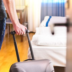 Person pulling suitcase, entering a hotel room with a made bed, wooden floor, and a chair in the background.