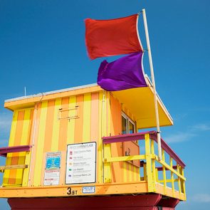 Colorful lifeguard tower with red and purple flags fluttering, situated on a beach under a clear blue sky. Signs display beach safety information.
