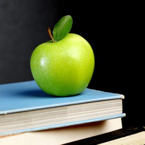 A green apple rests on stacked books against a dark background, with a single green leaf attached.