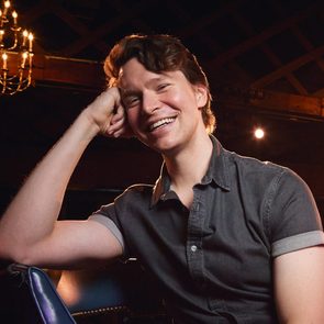 A person smiling, resting their head on their hand, seated indoors under a dimly lit chandelier.