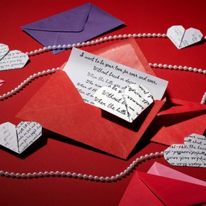 Envelope containing a handwritten note, surrounded by paper hearts and pearl strands on a red surface.