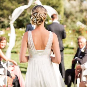 Bride walking down an aisle outdoors, guests watching and smiling, with a floral arch and greenery in the background.