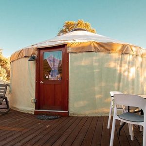Circular yurt stands on wooden deck, surrounded by chairs; features a wooden door with 