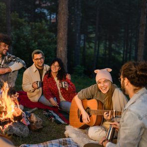 Group sits around campfire; one plays guitar. Forest setting, warm atmosphere, people smiling, holding mugs, enjoying a cozy evening.