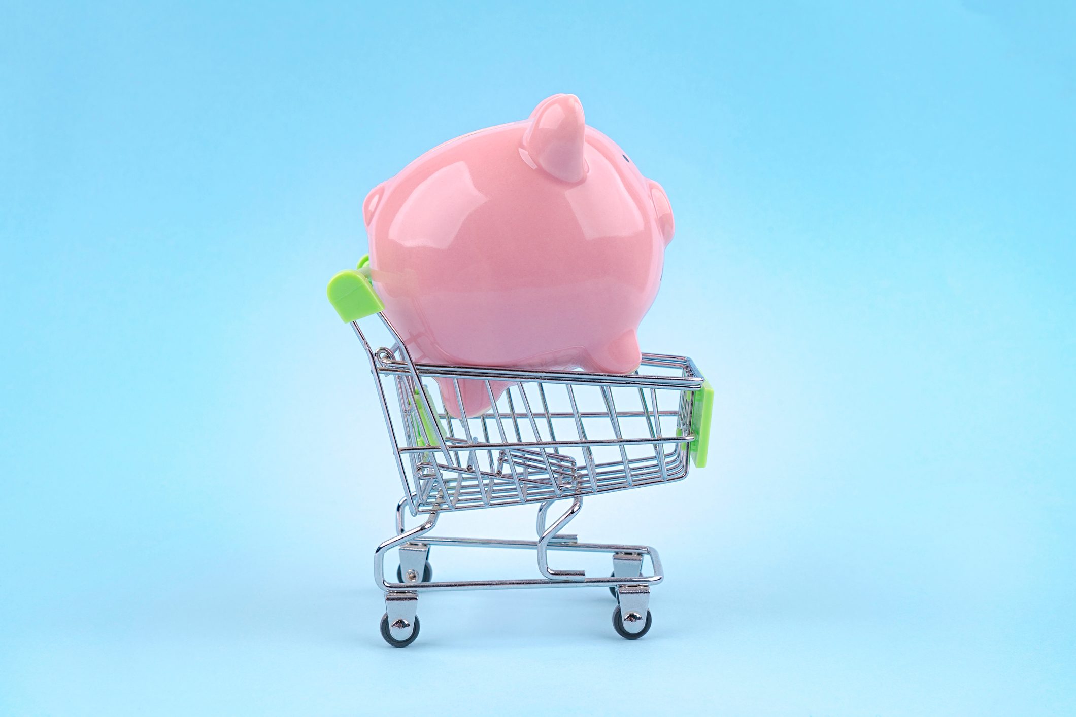 A pink piggy bank sits inside a small shopping cart against a plain blue background.