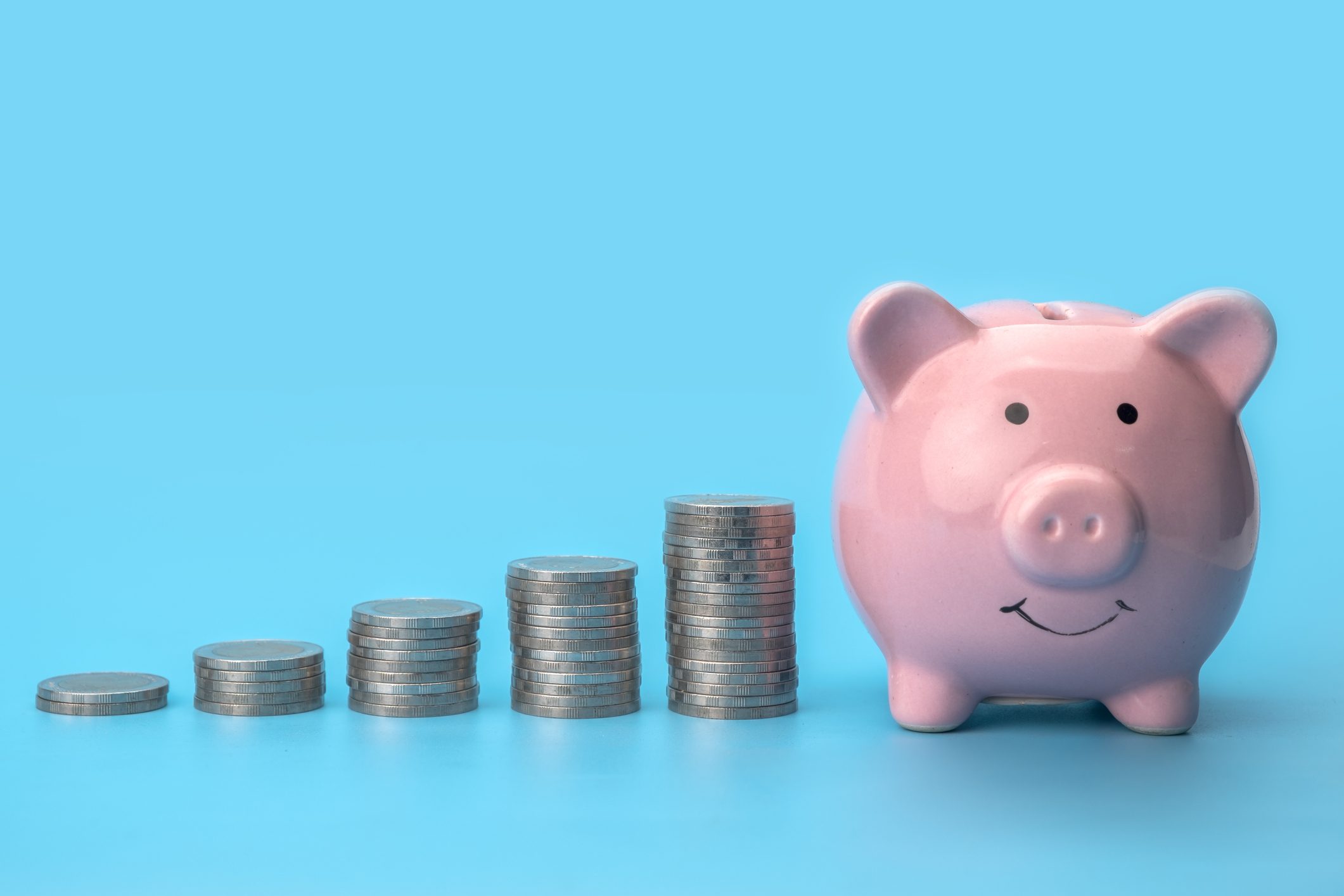 Piggy bank sitting beside increasing stacks of coins on a light blue background.