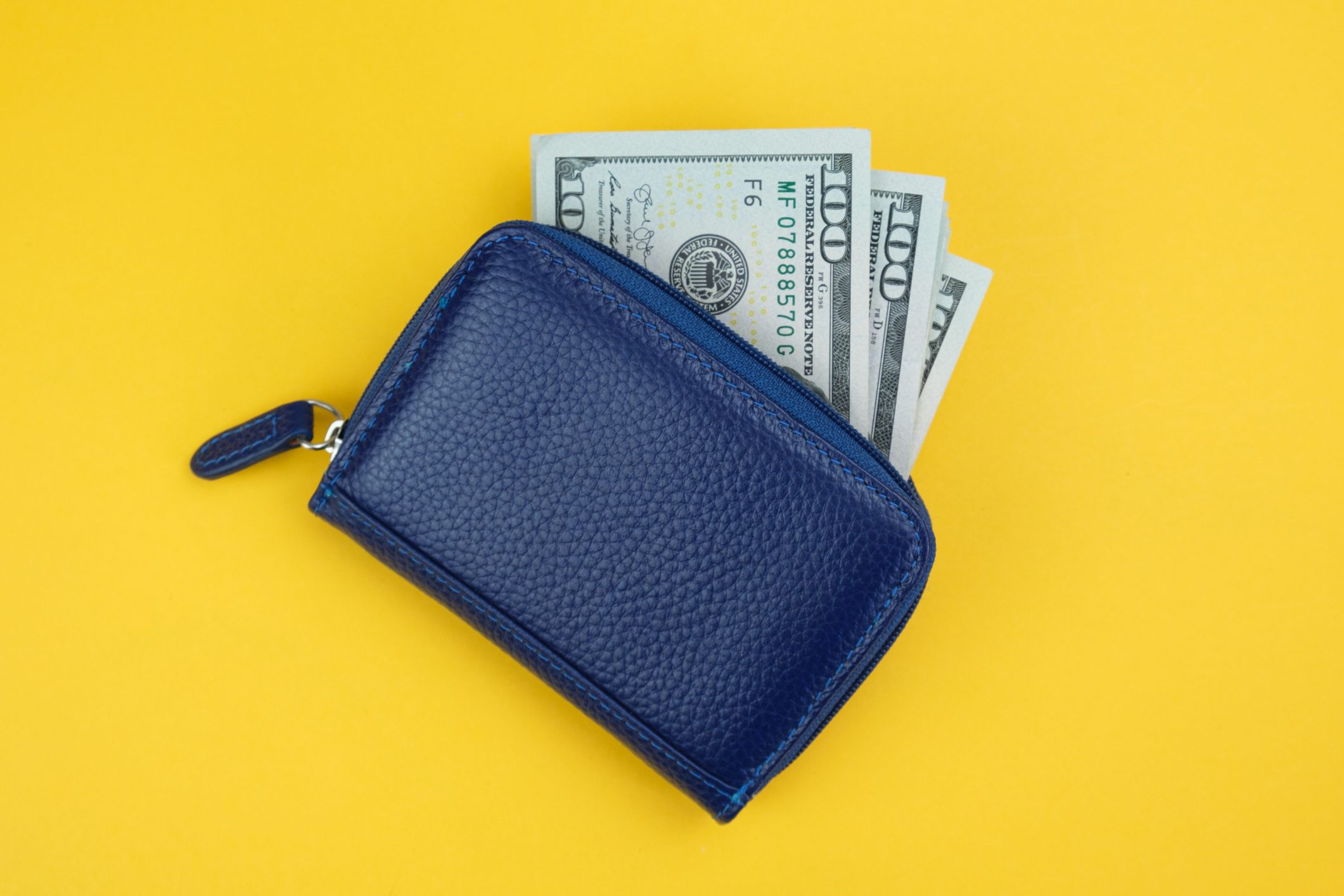 A blue wallet rests on yellow, containing visible U.S. hundred-dollar bills partially sticking out.