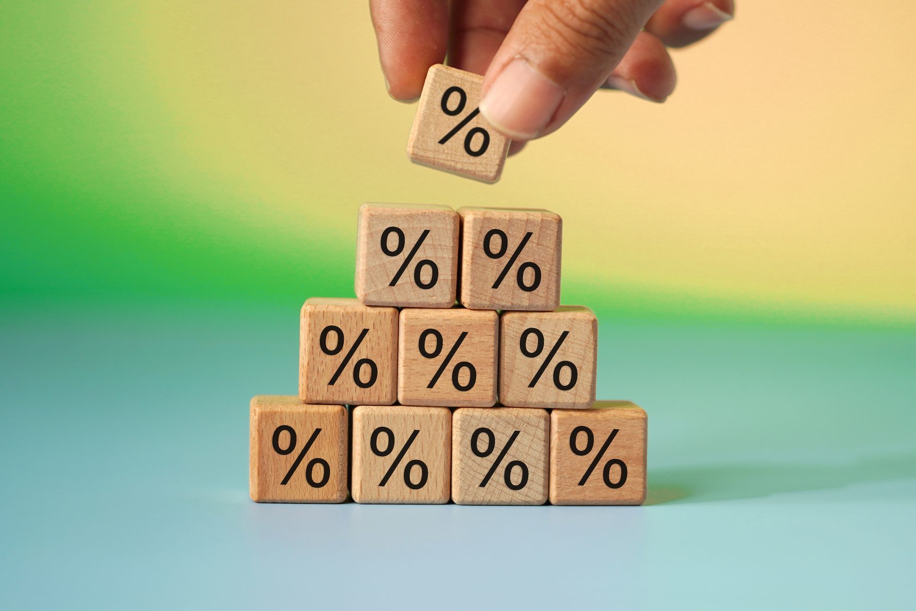 Hand stacks wooden blocks marked with percentage symbols in a pyramid shape on a colorful gradient background.