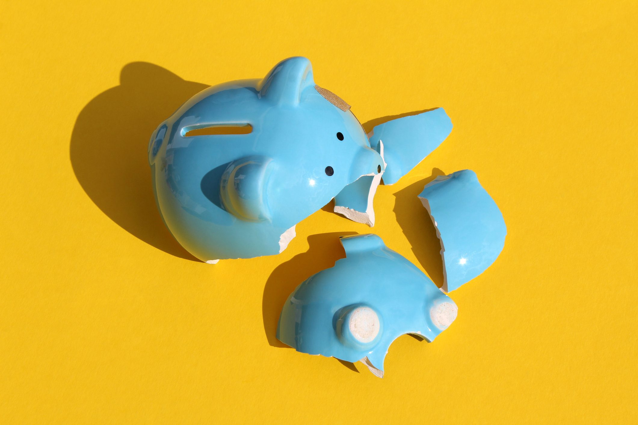 Broken blue piggy bank lies shattered across bright yellow background.