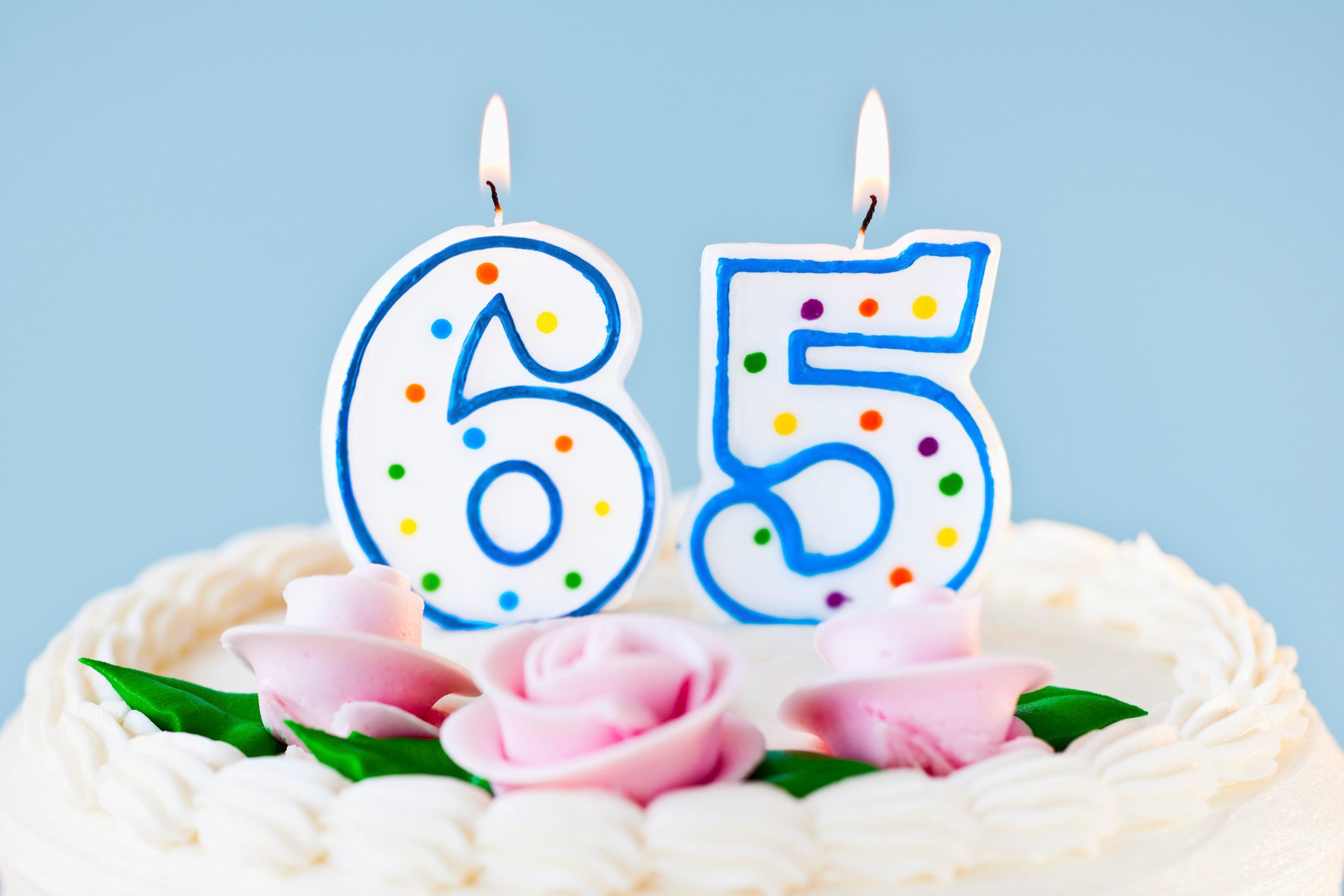 Candles depicting "65" are lit atop a cake, decorated with pink roses and icing, against a light blue background.