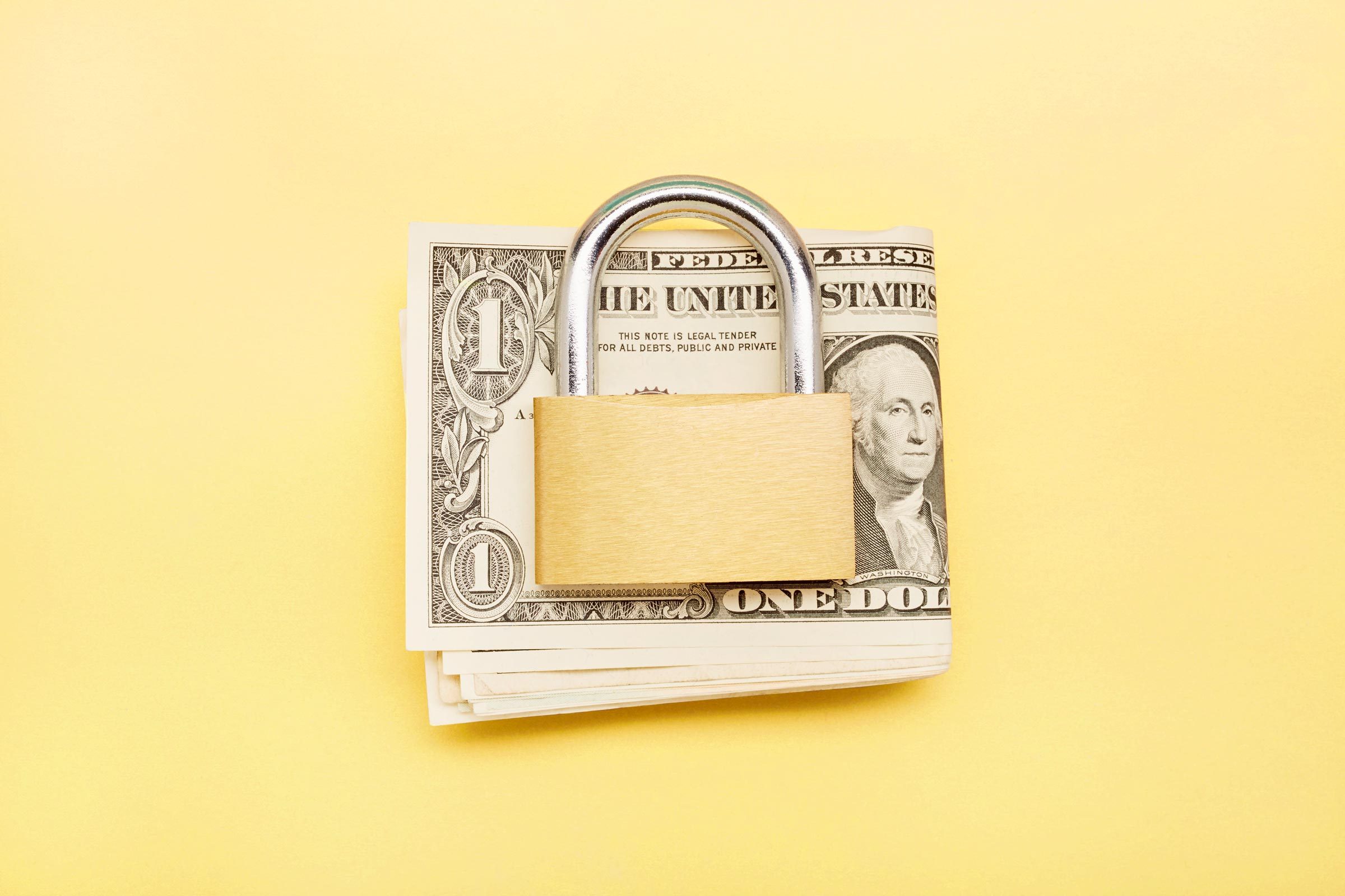 A padlock secures a stack of dollar bills against a smooth yellow background.